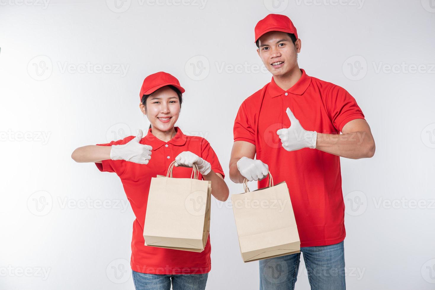 Bild eines glücklichen jungen Zustellers in roter Kappe, leerem T-Shirt, Uniform, stehend mit leerem braunem Kraftpapierpaket, isoliert auf hellgrauem Hintergrund Studio foto