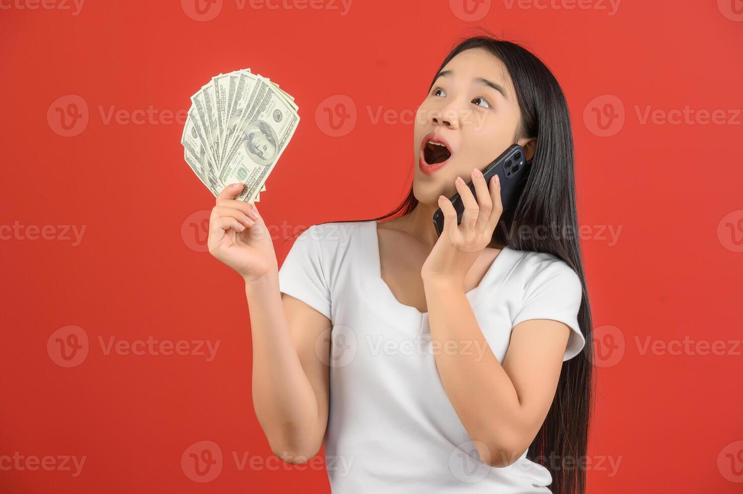 Porträt einer fröhlichen jungen Frau, die Geldscheine hält und mit dem Handy spricht, isoliert auf rotem Hintergrund foto