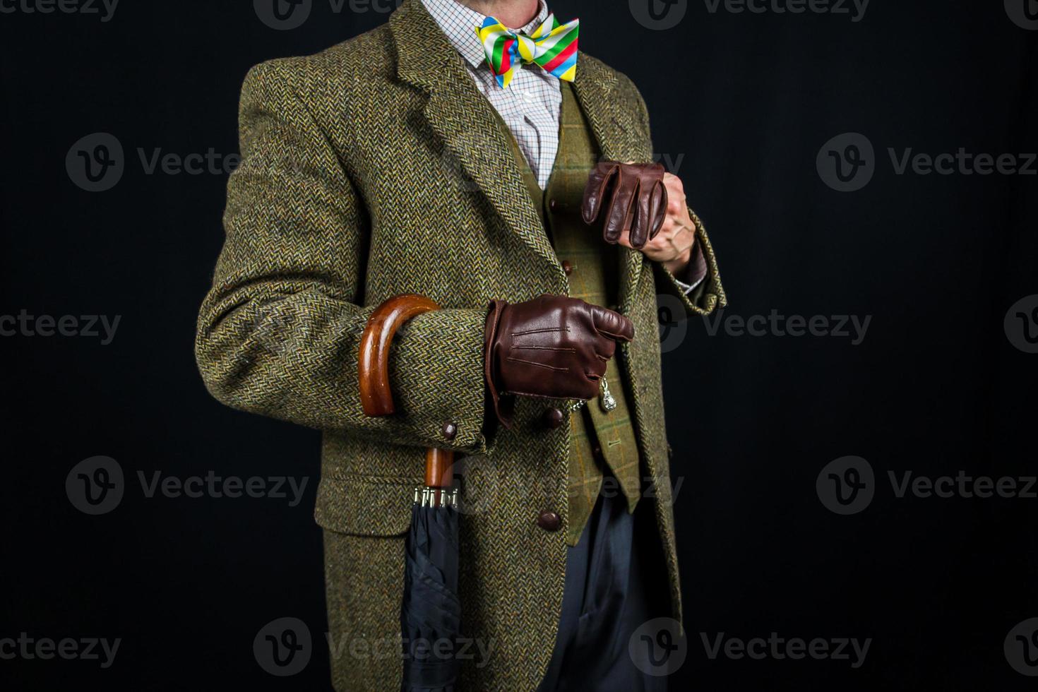 Gentleman in Tweed-Anzug und Lederhandschuhen mit Regenschirm am Arm auf schwarzem Hintergrund. Konzept des klassischen und exzentrischen britischen Gentleman-Stereotyps. Retro-Stil und Vintage-Mode. foto