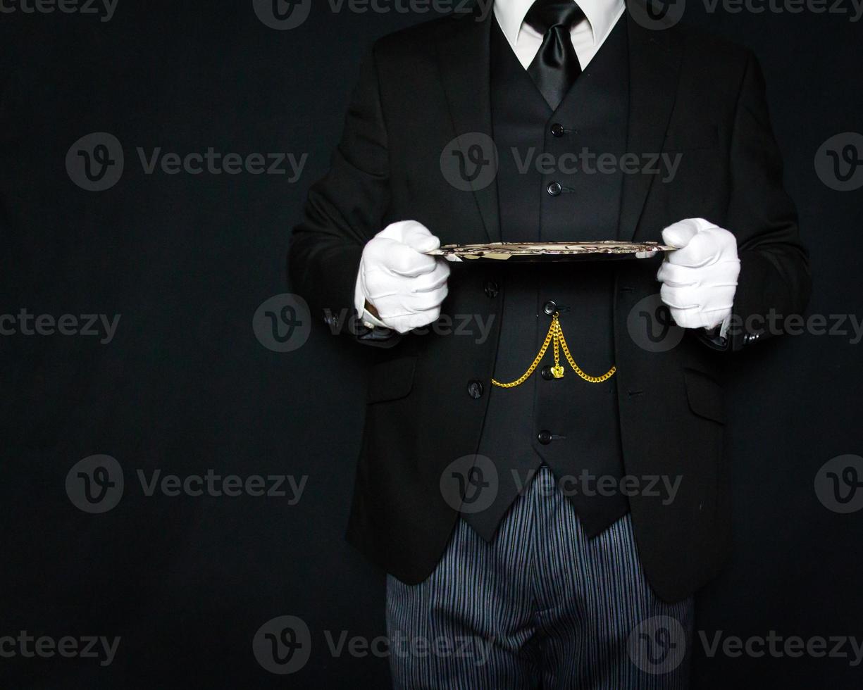 Porträt eines Butlers in dunklem Anzug und weißen Handschuhen, der ein silbernes Serviertablett auf schwarzem Hintergrund hält. kopierraum für die dienstleistungsbranche und professionelle gastronomie. foto