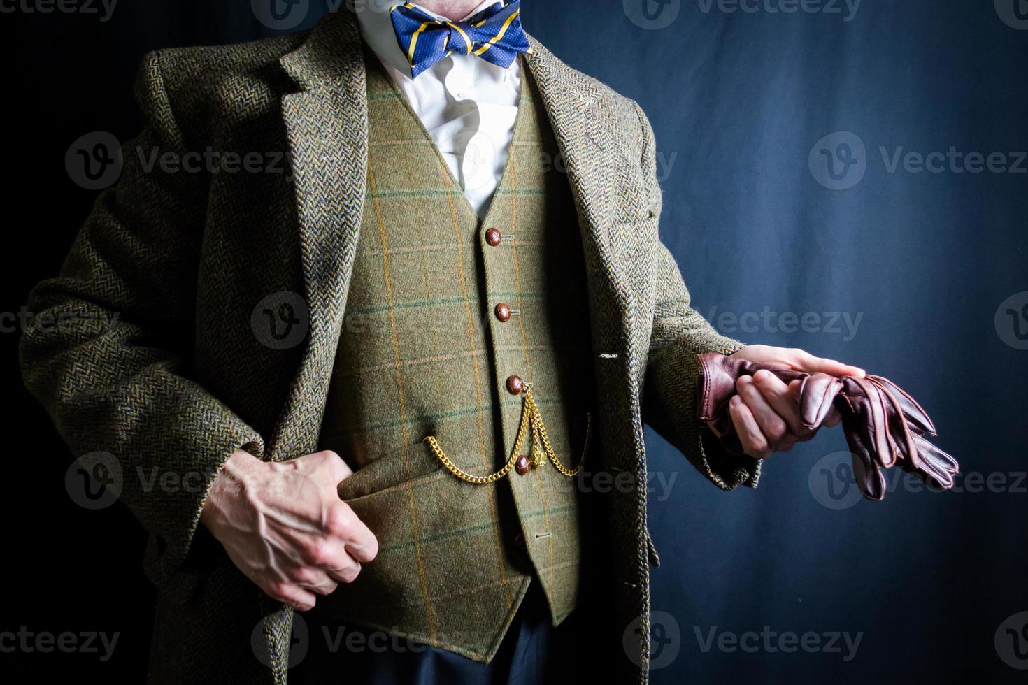 Porträt eines Mannes im Tweed-Anzug, der ein Paar braune Lederhandschuhe mit unbekümmerter Haltung auf schwarzem Hintergrund hält. klassischer und exzentrischer englischer Gentleman. foto