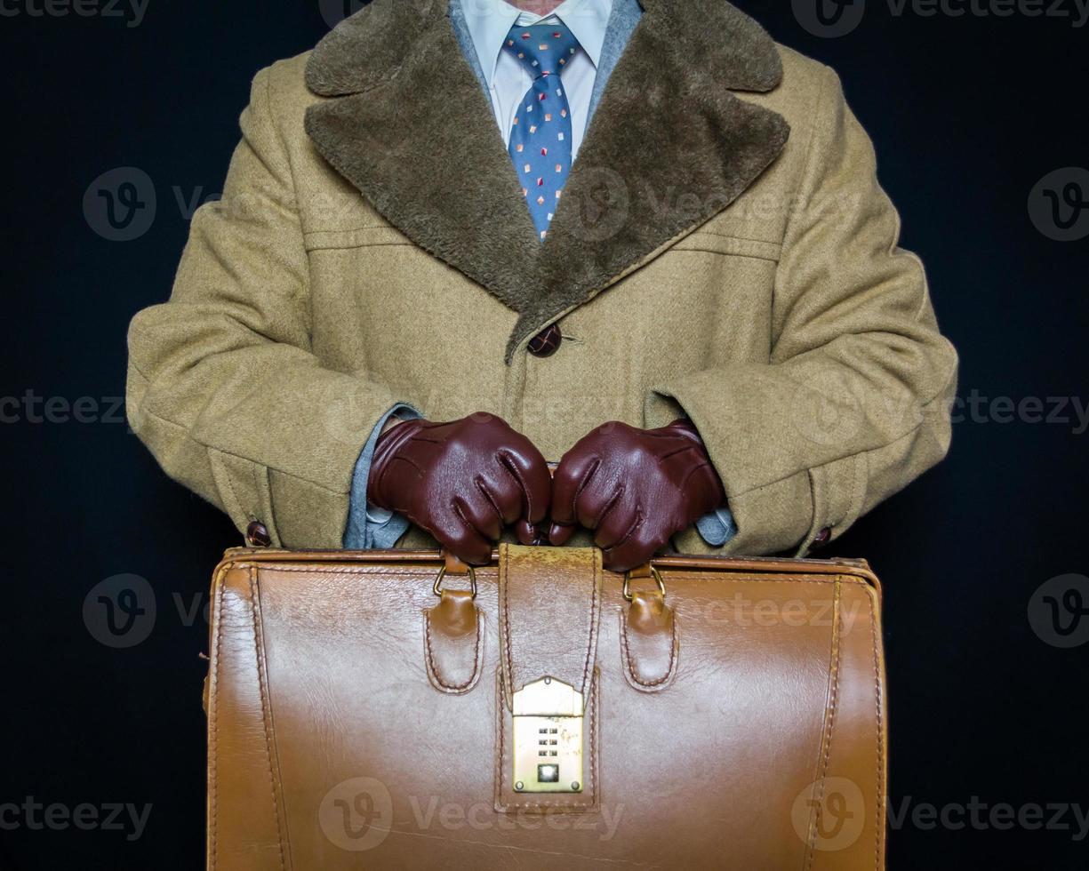Porträt eines Mannes im Wintermantel mit Vintage-Reisetasche auf schwarzem Hintergrund. Geheimagent film noir. foto