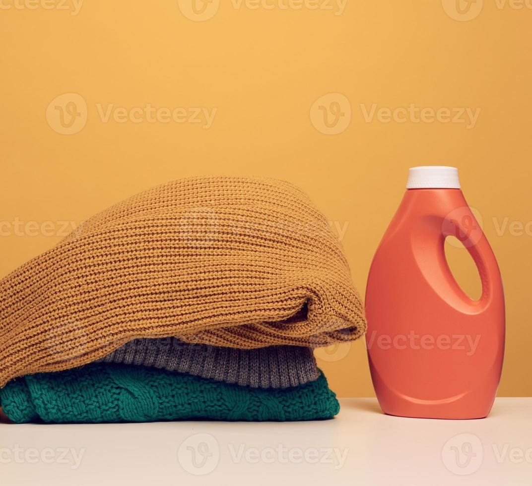 Stapel gewaschener gefalteter Kleidung und orange große Plastikflasche mit Flüssigwaschmittel stehen auf einem weißen Tisch, gelber Hintergrund. Routine Hausaufgaben foto
