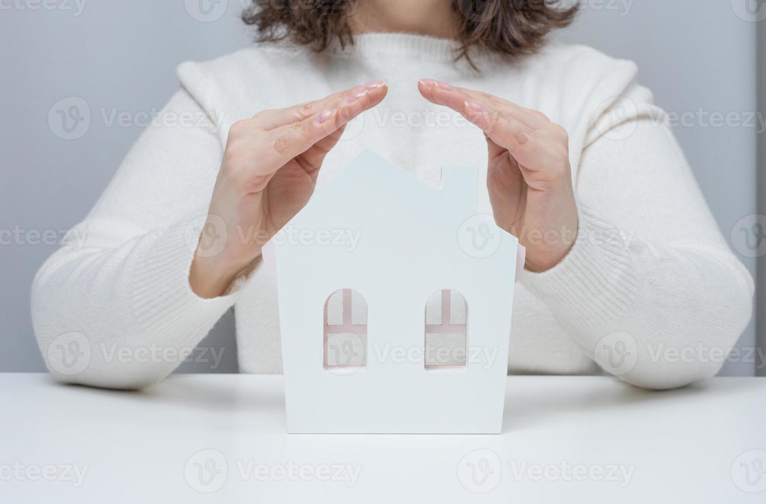 Weibliche Hände, die über einem hölzernen Miniaturmodellhaus auf weißem Hintergrund zusammengefaltet sind. immobilienversicherungskonzept, umweltschutz foto