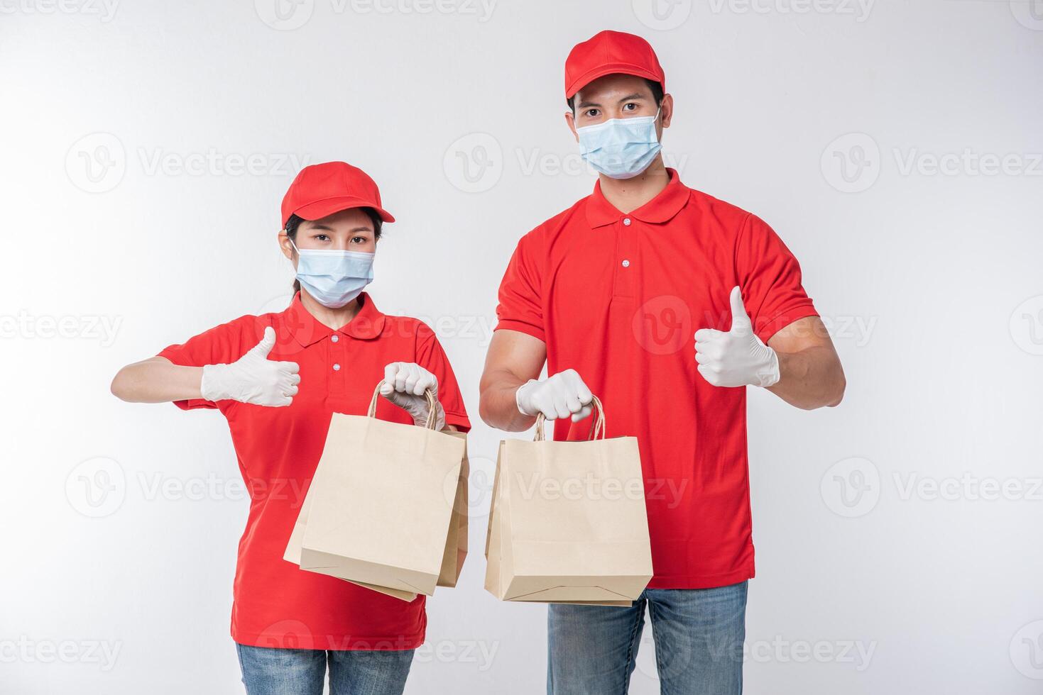 Bild eines glücklichen jungen Zustellers mit roter Kappe, leerem T-Shirt, einheitlichen Gesichtsmaskenhandschuhen, die mit leerem braunem Kraftpapierpaket stehen, das auf hellgrauem Hintergrundstudio isoliert ist foto