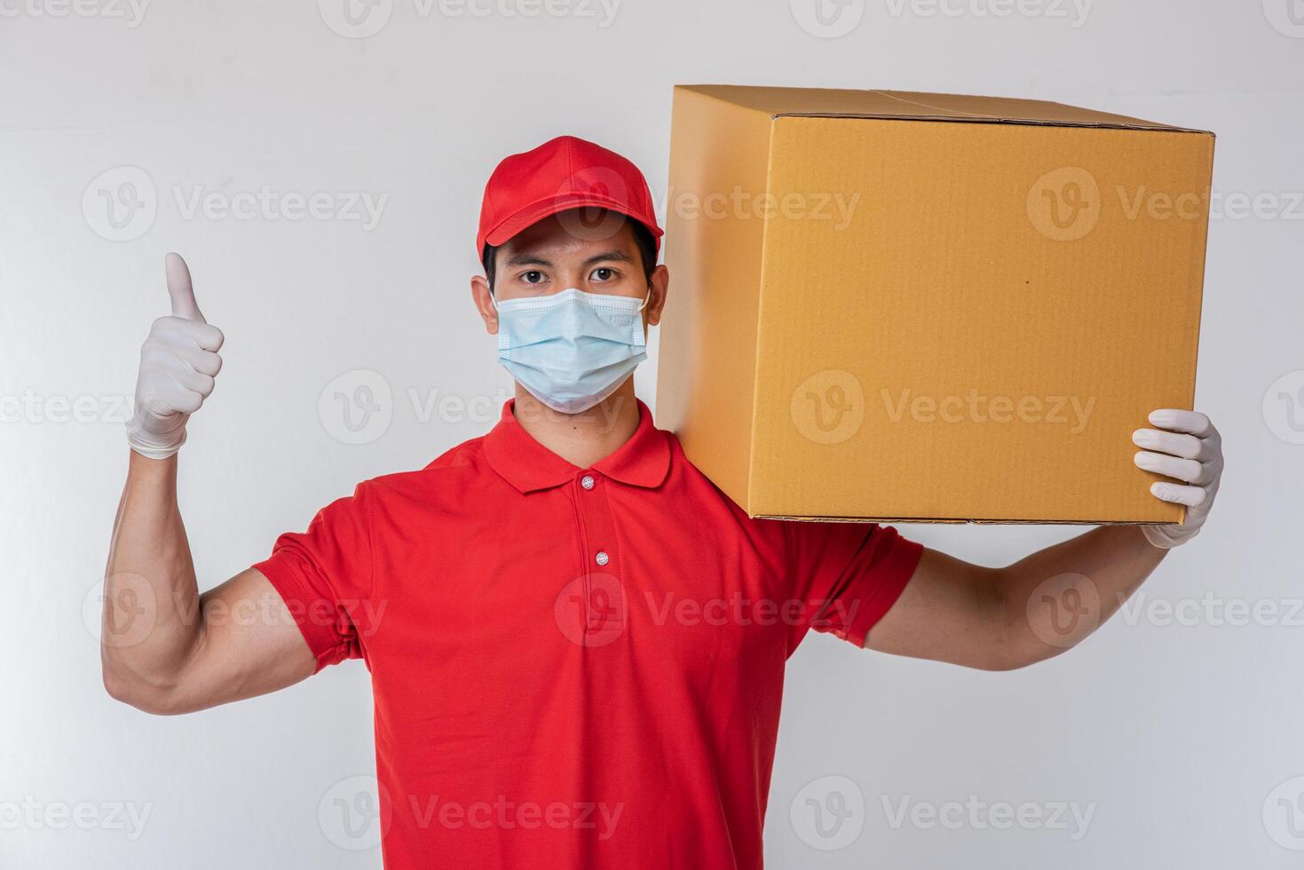 Bild eines bewussten jungen Zustellers mit roter Kappe, leerem T-Shirt, einheitlichen Gesichtsmaskenhandschuhen, die mit leerem braunem Karton stehen, isoliert auf hellgrauem Hintergrund Studio foto