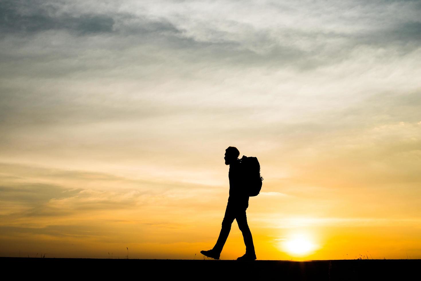 Schattenbild eines jungen Rucksacktouristenmannes, der während des Sonnenuntergangs geht foto