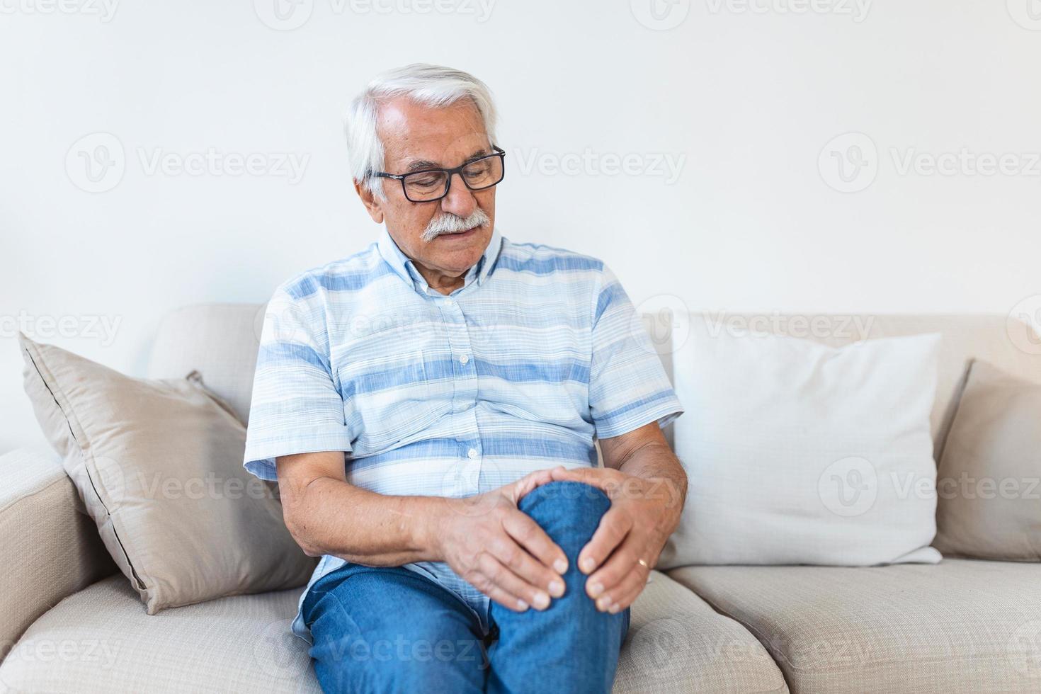 älterer Mann, der zu Hause auf einem Sofa sitzt und sein schmerzhaftes Knie berührt. menschen, gesundheitswesen und problemkonzept - unglücklicher senior, der zu hause unter knieschmerzen leidet foto