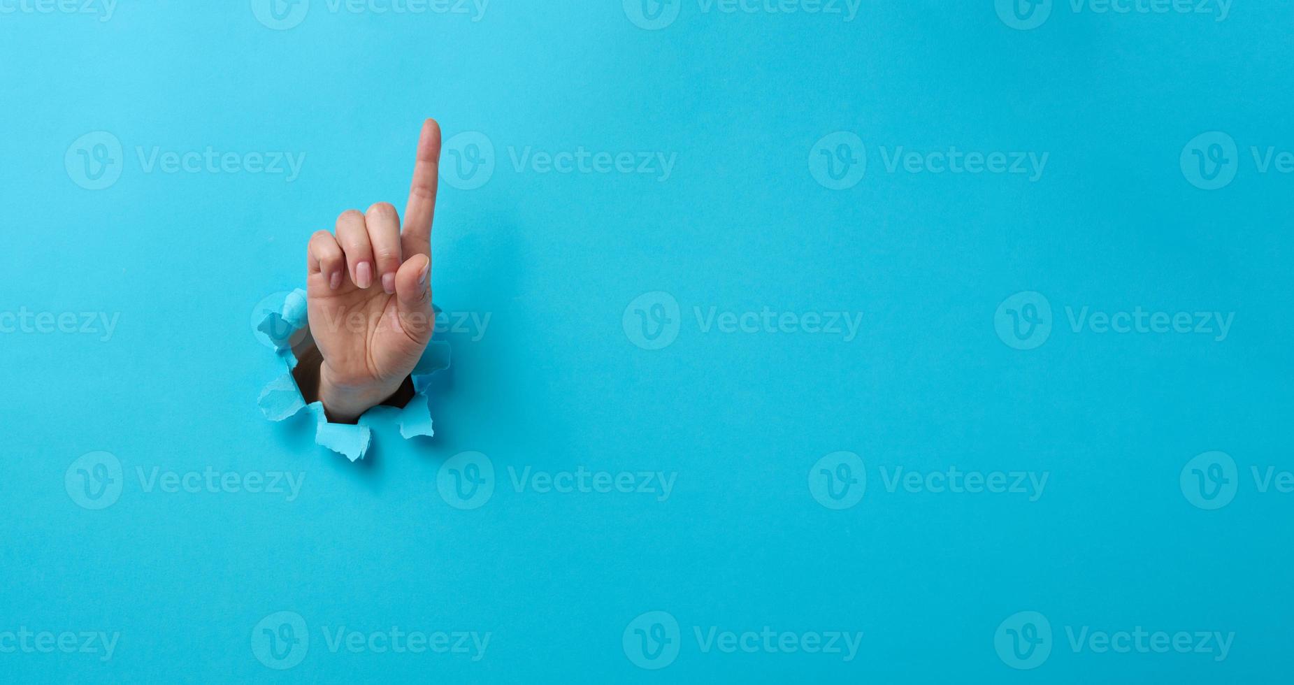 weiblich Hand kleben aus von ein zerrissen Loch im ein Blau Papier Hintergrund, Beachtung Geste. Platz zum ein Inschrift, Banner foto