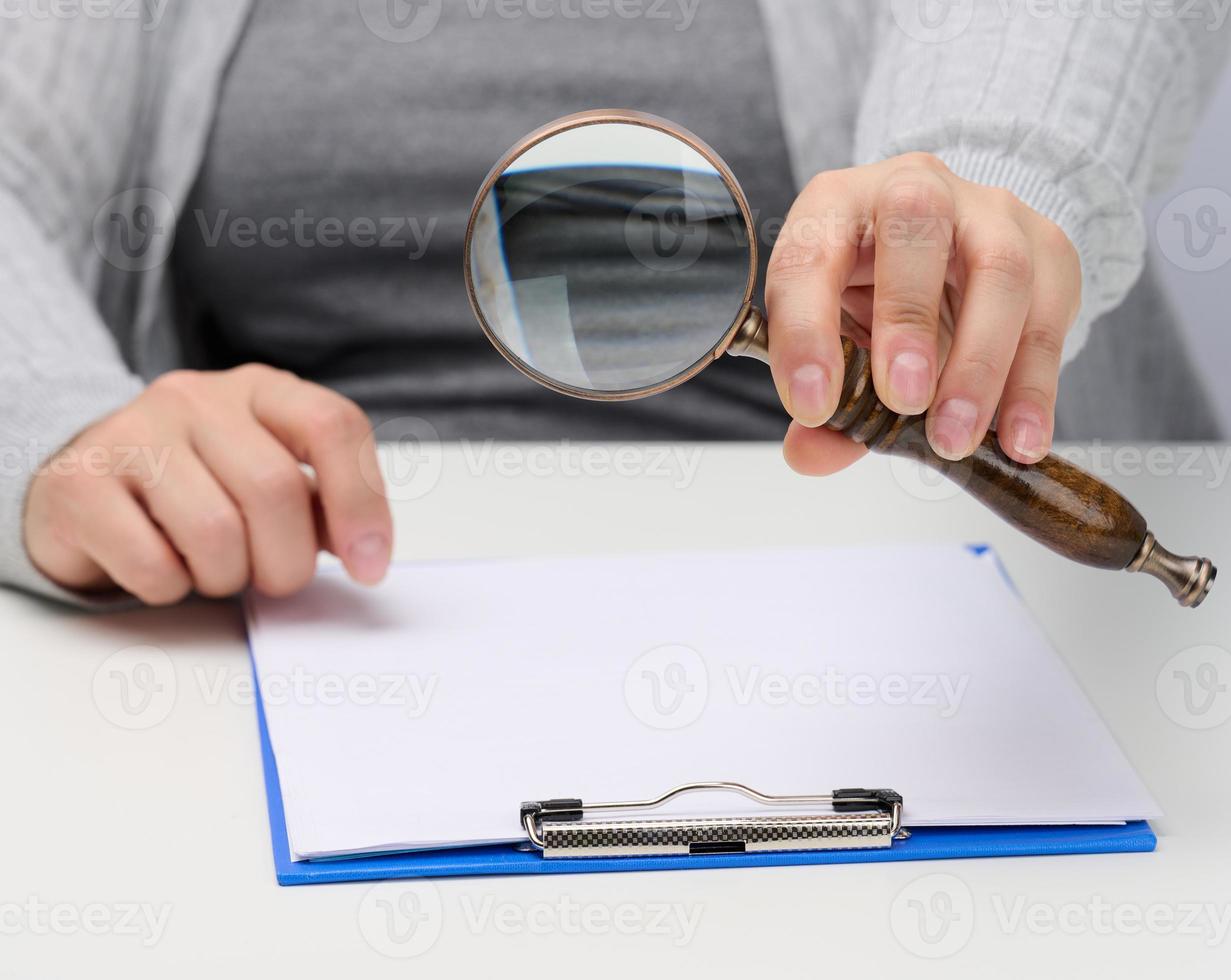 weiblich Hand hält ein hölzern Vergrößerung Glas Über ein Weiß Tabelle mit Unterlagen. Suche zum Antworten zu Fragen, Geschäft Analyse foto
