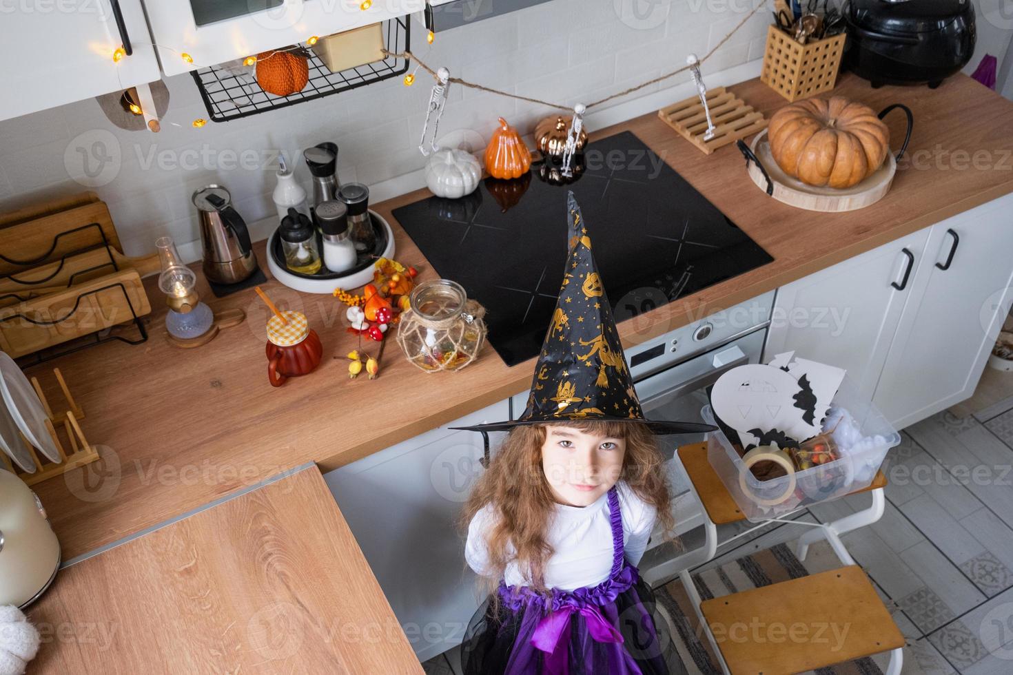 Kind schmückt die Küche zu Hause für Halloween. Mädchen in einem Hexenkostüm spielt mit dem Dekor für den Urlaub - Fledermäuse, Jack-Laterne, Kürbisse. Herbstkomfort im Haus, Küche im Scandi-Stil, Dachboden foto