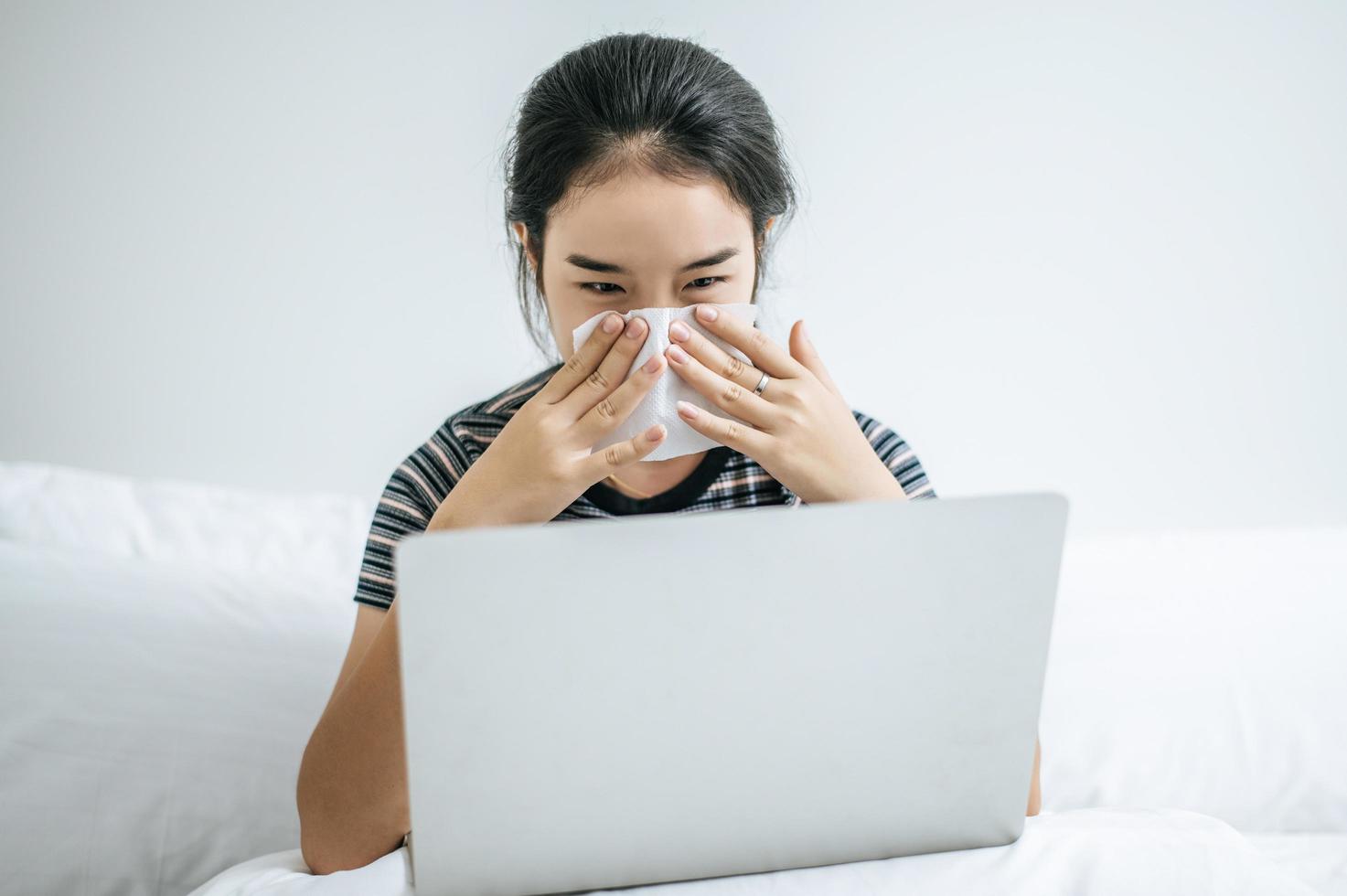 Frau spielt auf Laptop hält ein Taschentuch, um Nase zu wischen foto