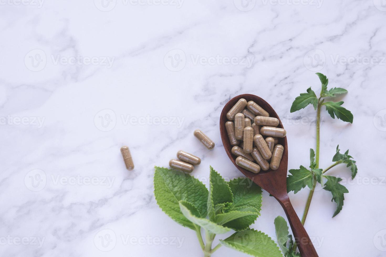 Kräutermedizin-Kapseln auf Holzlöffel mit grünem Blatt auf weißem Marmor foto