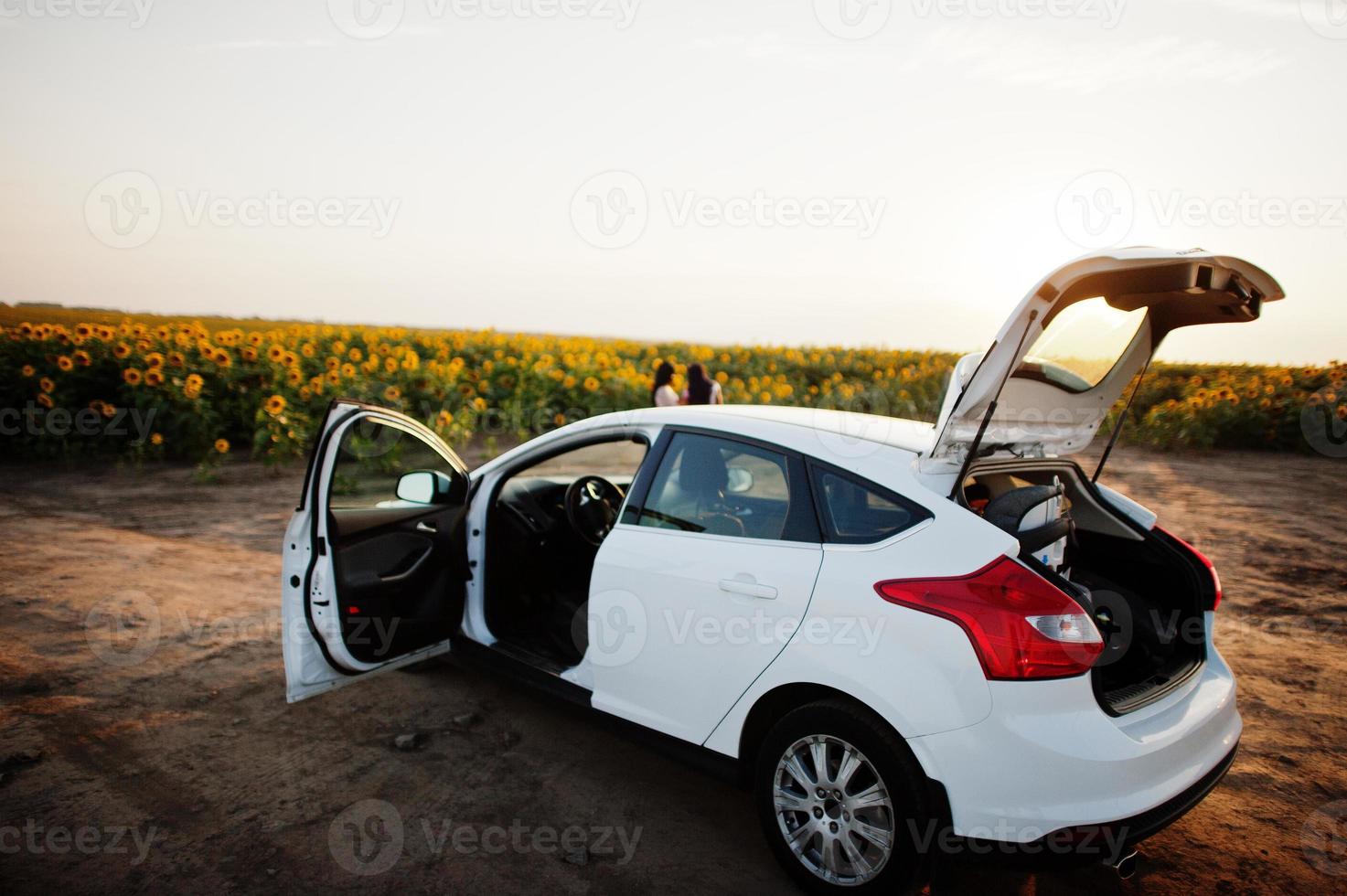 Weißes Auto gegen zwei hübsche schwarze Frauen tragen Sommerkleider in einem Sonnenblumenfeld. foto