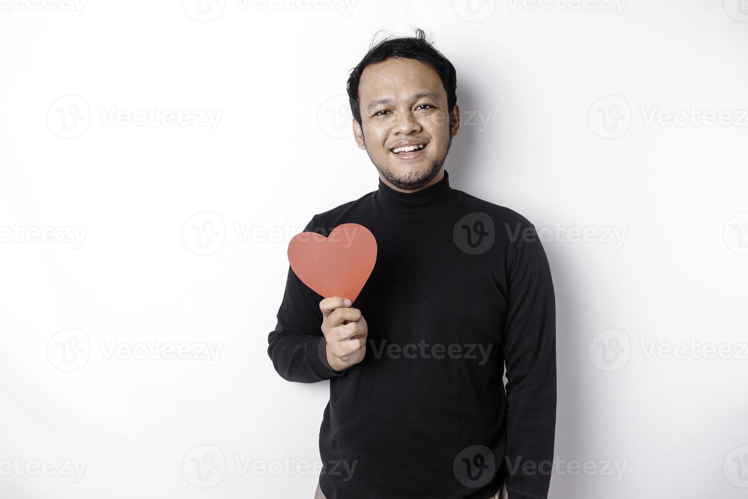 ein Porträt von ein glücklich asiatisch Mann tragen ein schwarz Shirt, halten ein rot herzförmig Papier isoliert durch Weiß Hintergrund foto