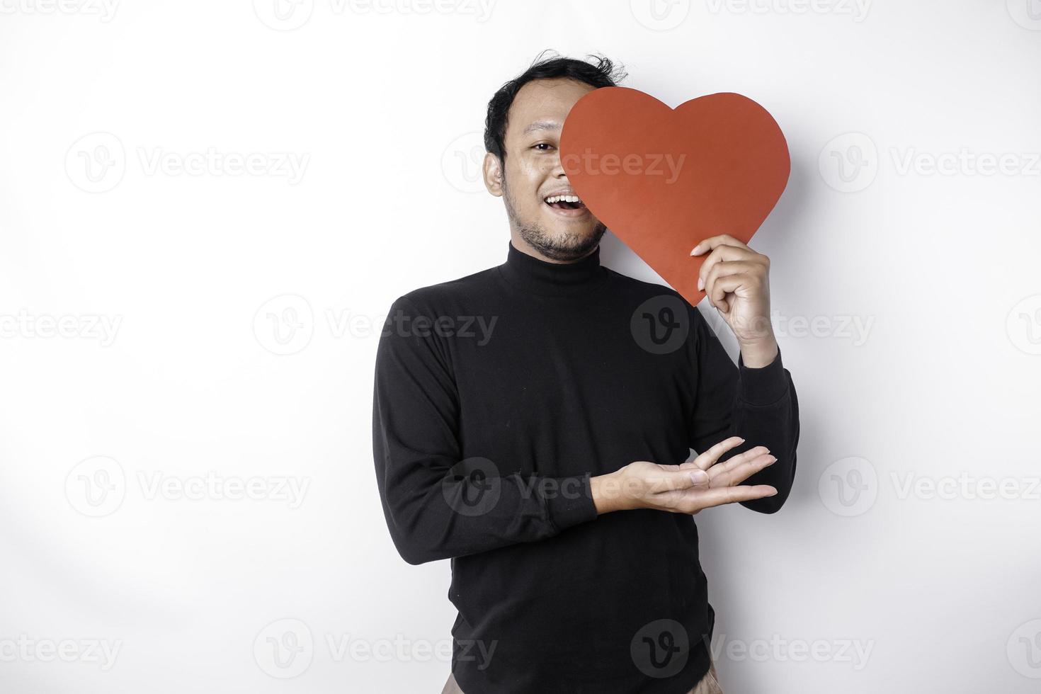 aufgeregt asiatisch Mann tragen schwarz Shirt, zeigen beim das Kopieren Raum neben ihm während halten ein groß rot herzförmig Papier, isoliert durch Weiß Hintergrund foto