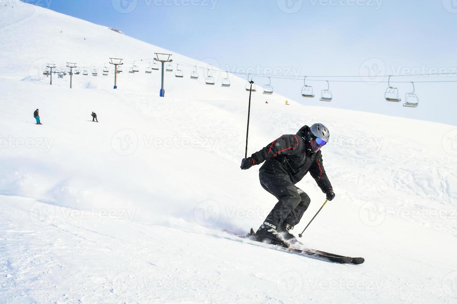 cool männlich Skifahrer im schwarz schließen oben Seite Aussicht gehen bergab Carving schnell Bewegung sprühen Schnee im bedeckt Tag mit Berge und Ski Aufzug Hintergrund foto