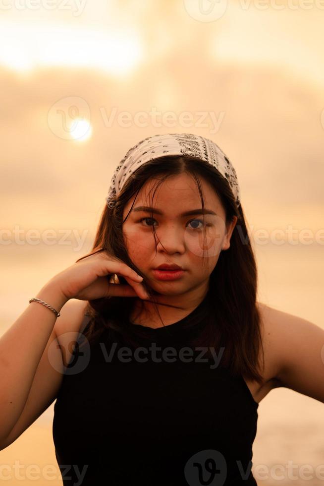 ein asiatisch Teenager tragen ein Weiß Bandana und schwarz Hemd mit ein eben Ausdruck während posieren mit ihr Haar auf das Strand foto