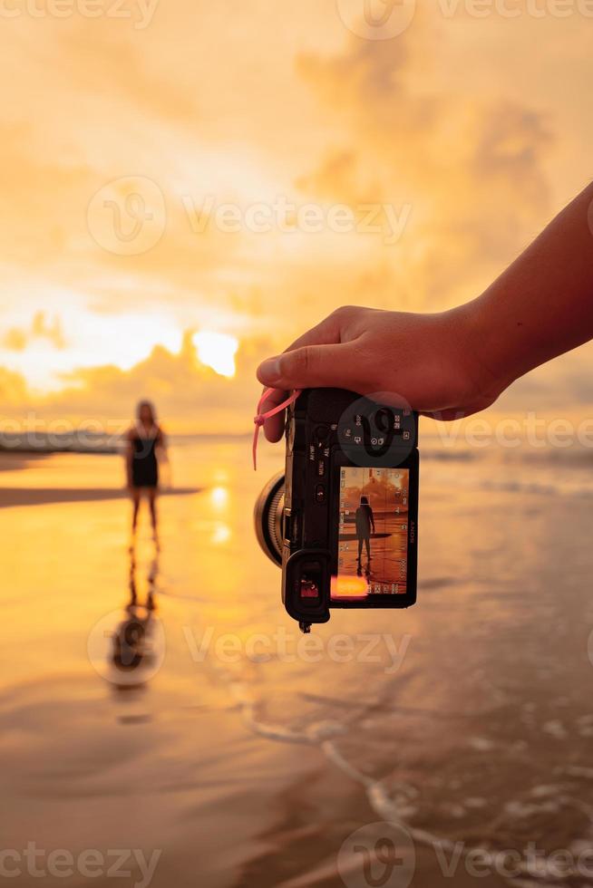 ein Kamera mit Hände Fotografien ein balinesisch Frau tun ein Gymnastik- Bewegung auf ein schwarz Hemd in der Nähe von das Strand foto