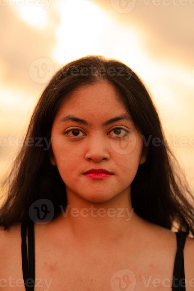 ein asiatisch Frau im ein schwarz Hemd mit ein Ausdruck Das sieht aus genervt ist Stehen in der Nähe von das Wellen auf das Strand im ein Urlaub foto