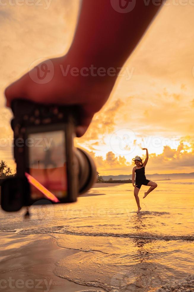 ein Kamera mit Hände Fotografien ein balinesisch Frau tun ein Gymnastik- Bewegung auf ein schwarz Hemd in der Nähe von das Strand foto