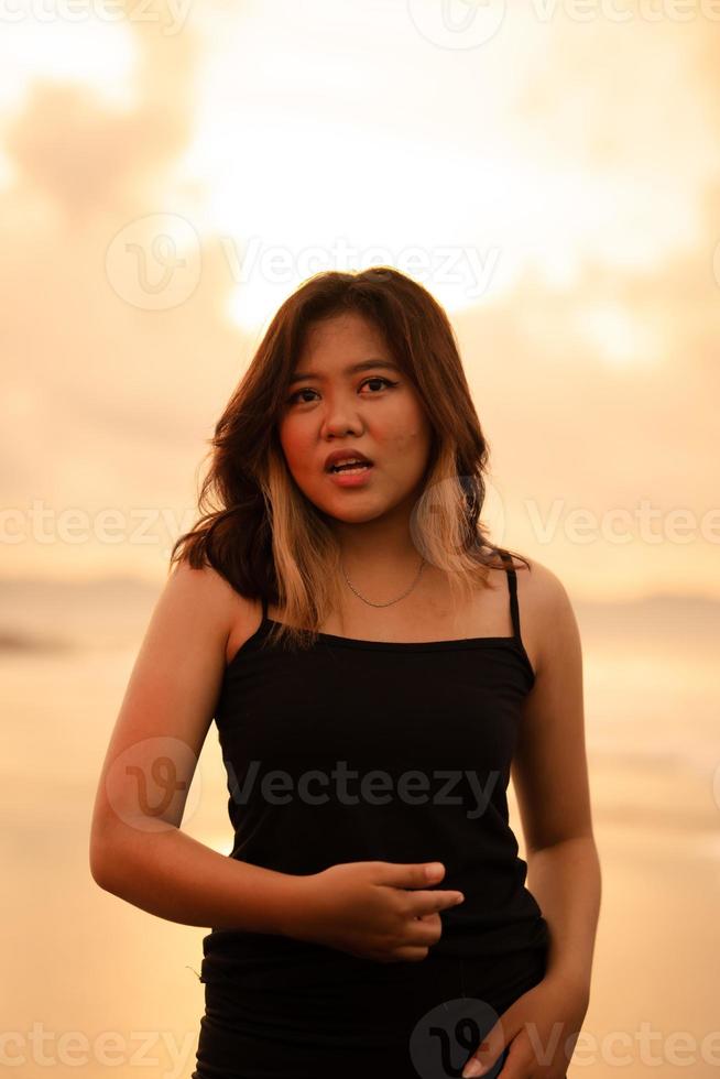 ein asiatisch Frau posiert mit ein schmutzig und wütend Ausdruck wann tragen ein schwarz Kleid auf das Strand foto