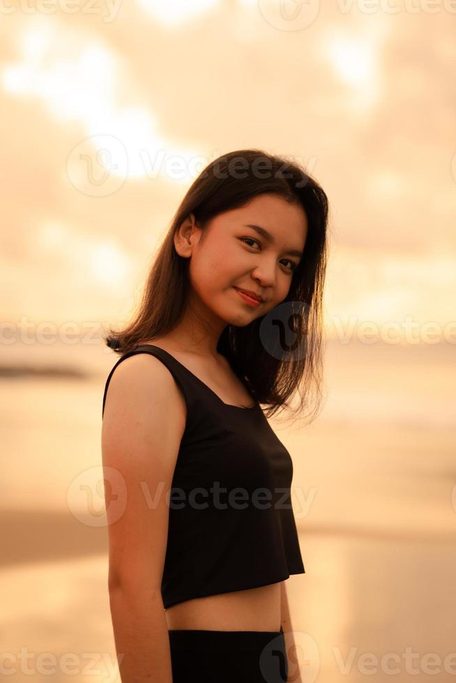 ein schön asiatisch Frau im schwarz Kleider ist genießen das Schönheit von das Strand mit ein lächelnd Ausdruck auf ihr Gesicht während auf Ferien foto