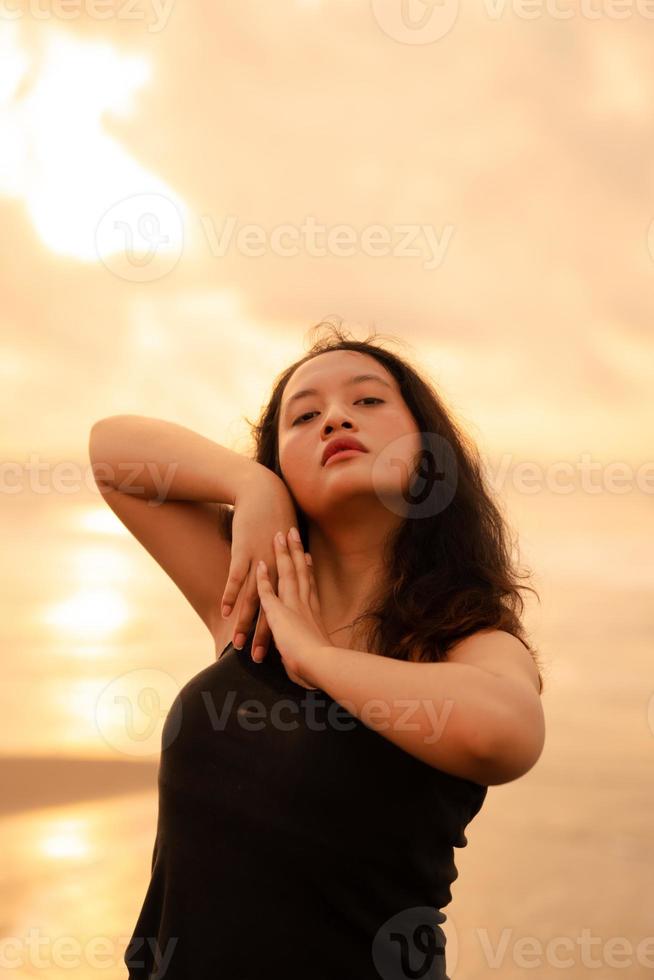 ein asiatisch Frau im ein schwarz Kleid und rundlich Körper Streiks ein sehr kokett Pose während genießen das Strand foto