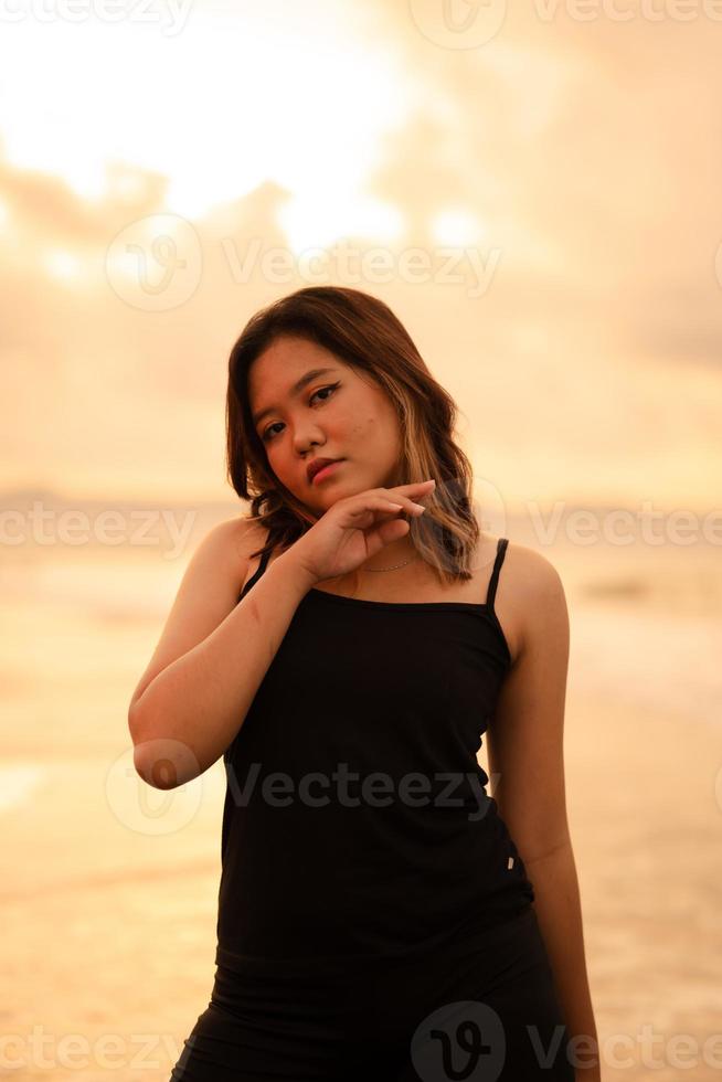 ein asiatisch Frau mit braun Haar und schwarz Kleider macht sexy posiert sehr geschickt während genießen das Strand foto