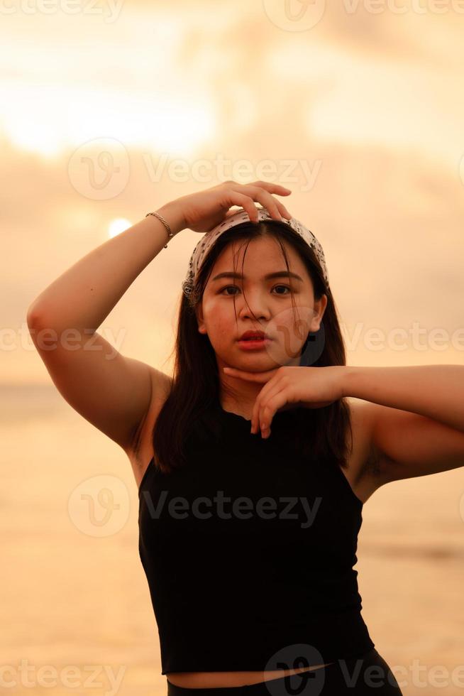 ein glamourös asiatisch Frau im schwarz Kleider und ein Weiß Bandana Stehen im Vorderseite von das Wellen auf das Strand foto