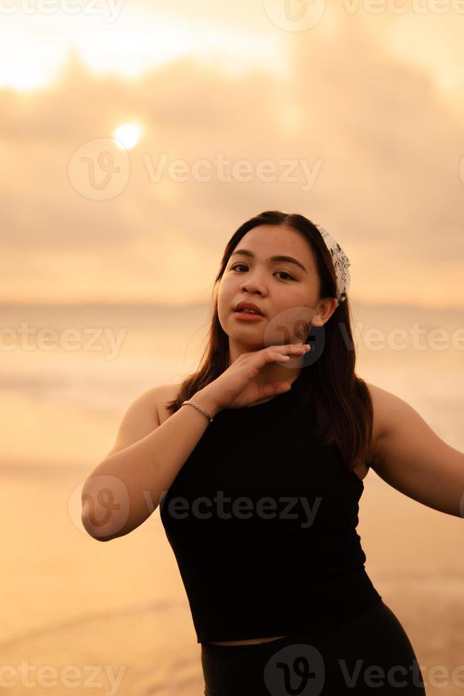 ein glamourös asiatisch Frau im schwarz Kleider und ein Weiß Bandana Stehen im Vorderseite von das Wellen auf das Strand foto