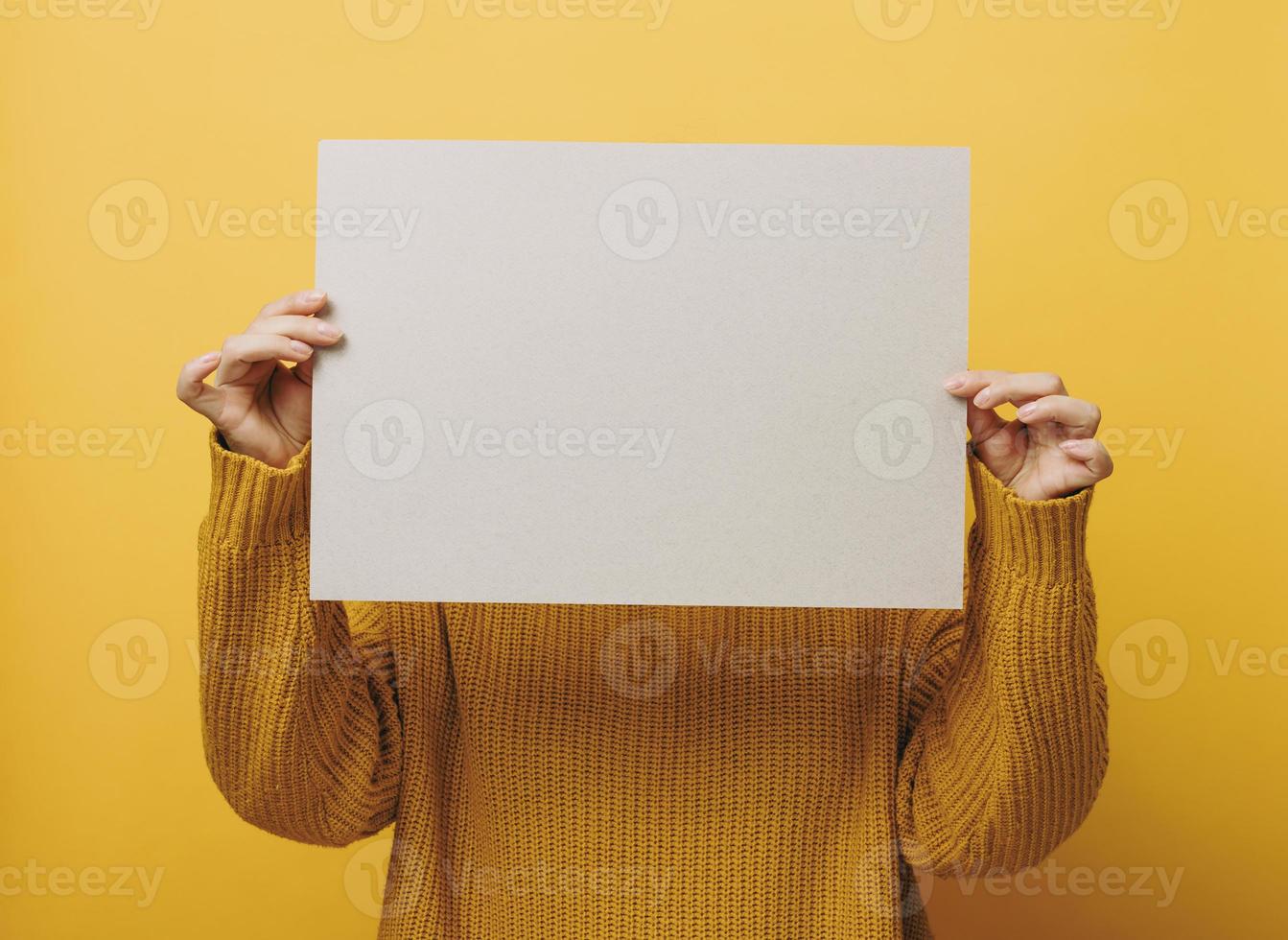Frau in einem orangefarbenen Pullover hält ein leeres Blatt Papier auf gelbem Hintergrund. platz für eine beschriftung, werbung, information foto