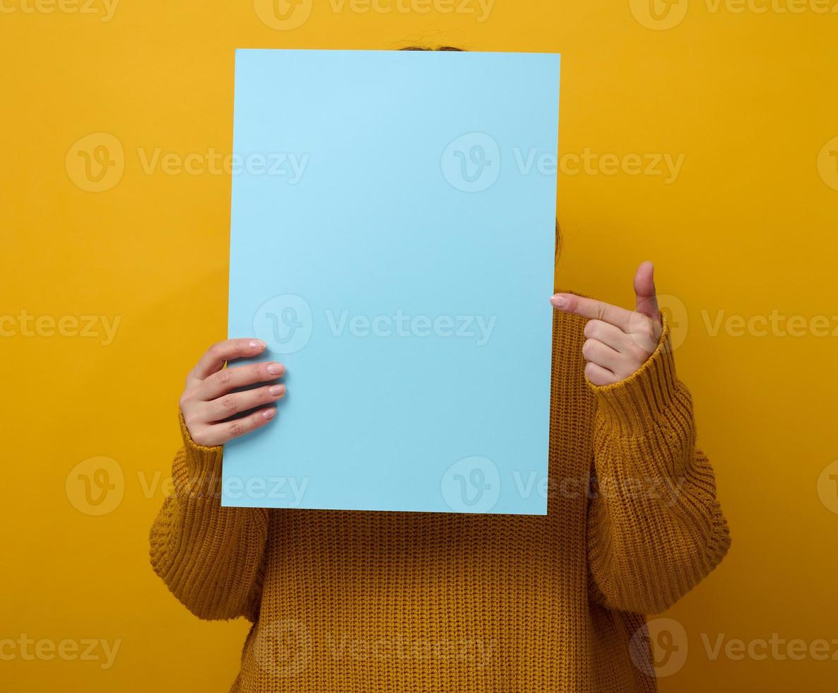 Frau in einem orangefarbenen Pullover hält ein leeres Blatt Papier auf gelbem Hintergrund. platz für eine beschriftung, werbung, information foto