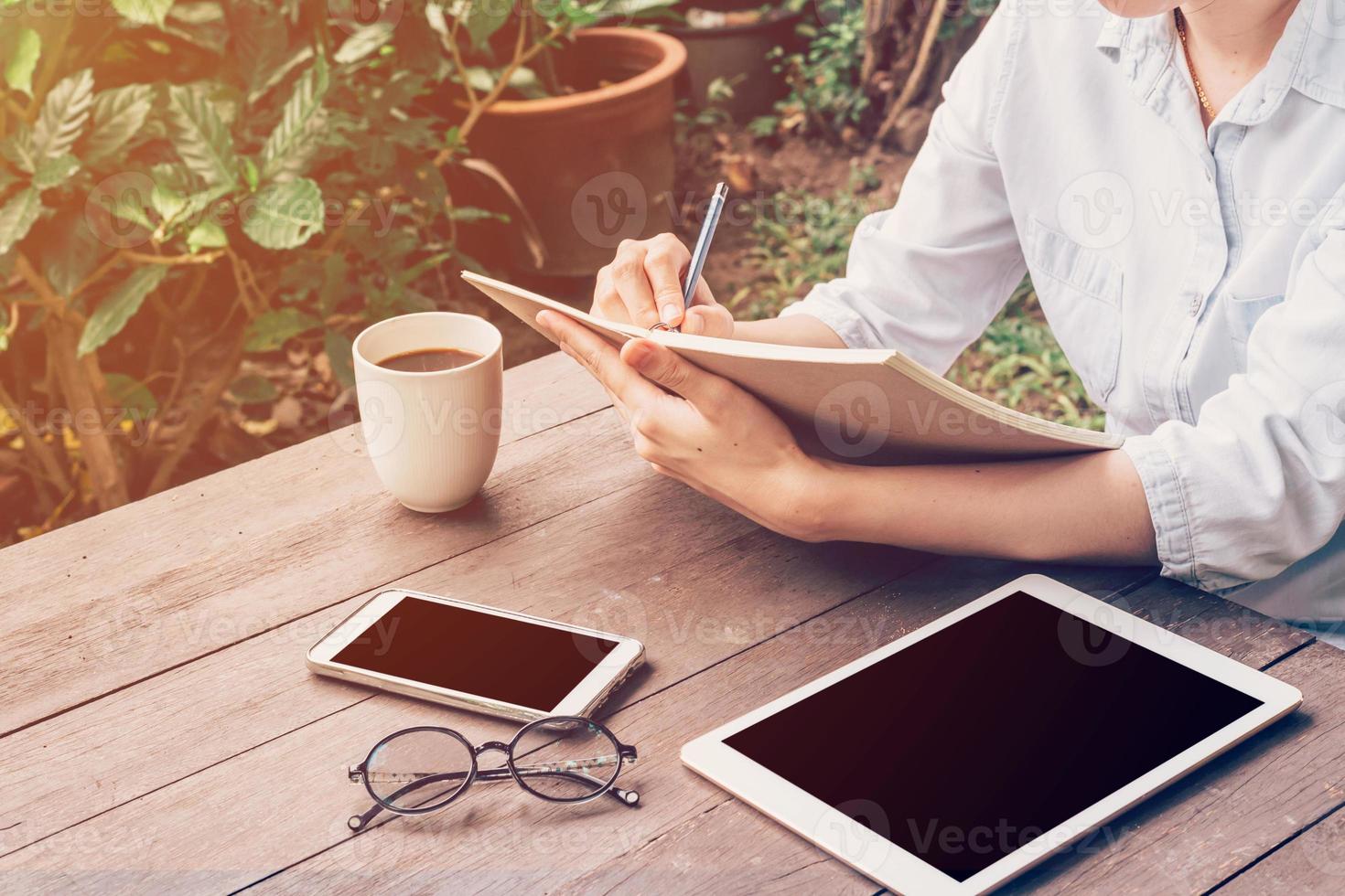 Frau Hand Schreiben Buch und Telefon, Tablette auf Tabelle im Garten beim Kaffee Geschäft mit Jahrgang getönt. foto