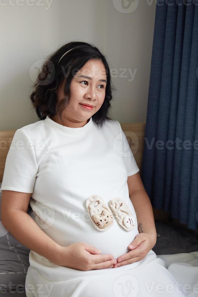 Foto von glücklich asiatisch schwanger Frau Sitzung im Bett und berühren ihr Bauch beim Zuhause mit klein Schuhe zum das ungeboren Baby im das Bauch