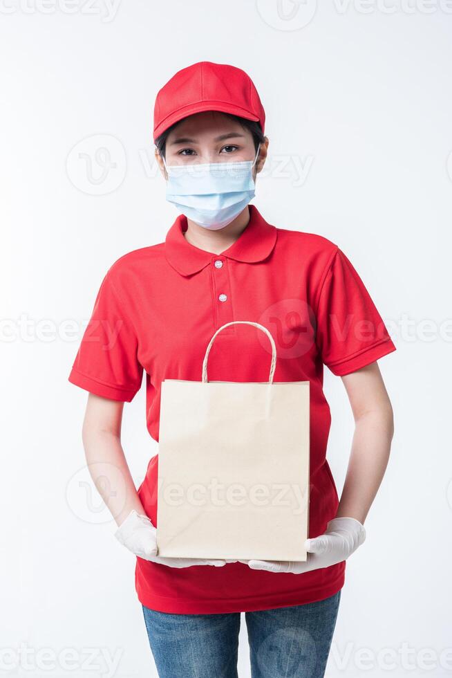 Bild eines glücklichen jungen Zustellers mit roter Kappe, leerem T-Shirt, einheitlichen Gesichtsmaskenhandschuhen, die mit leerem braunem Kraftpapierpaket stehen, das auf hellgrauem Hintergrundstudio isoliert ist foto