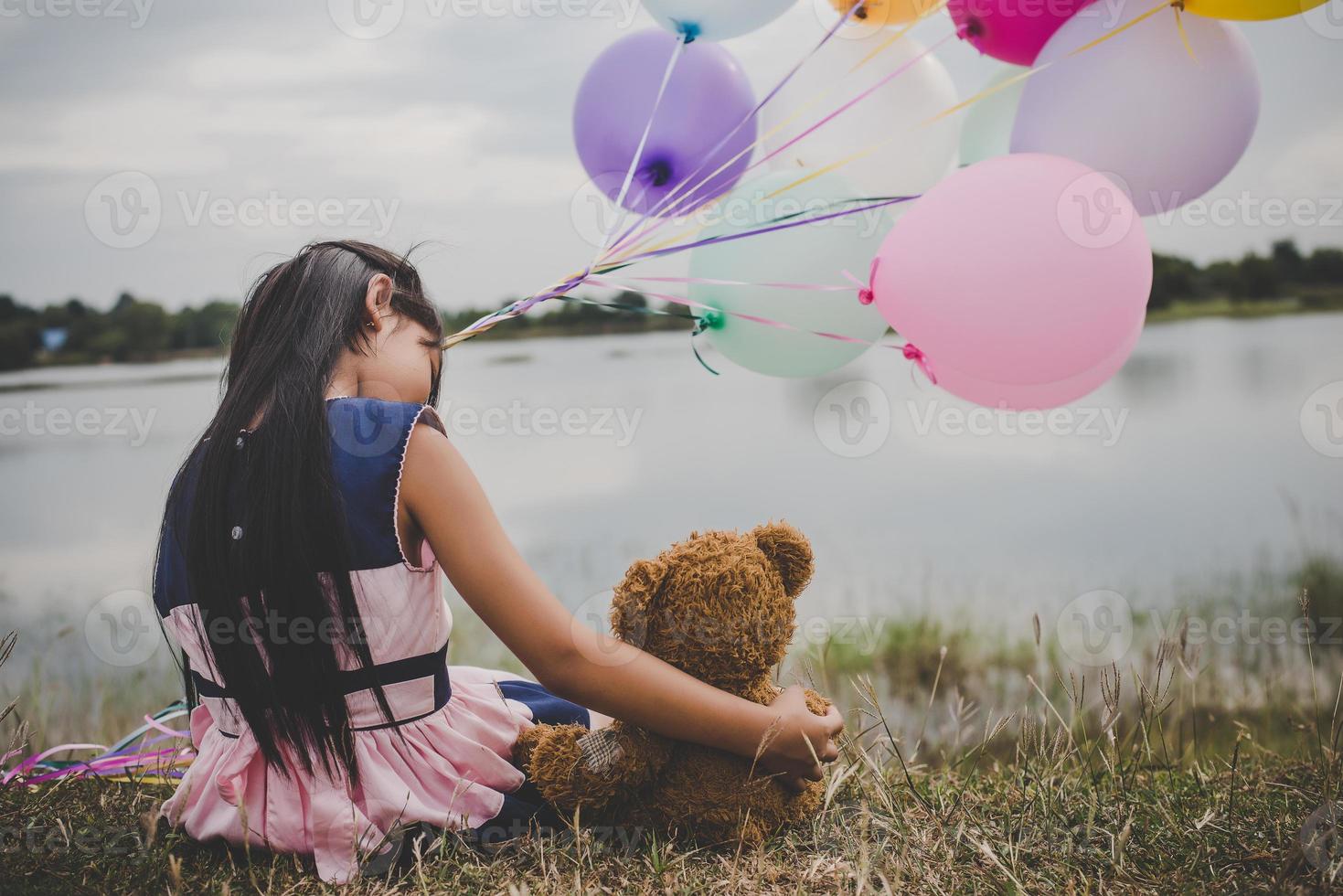 kleines Mädchen mit einem Teddybär und Luftballons auf Wiesenfeld foto