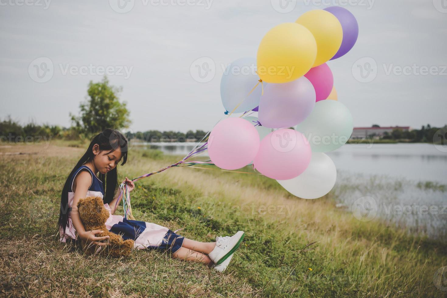 kleines Mädchen mit einem Teddybär und Luftballons auf Wiesenfeld foto