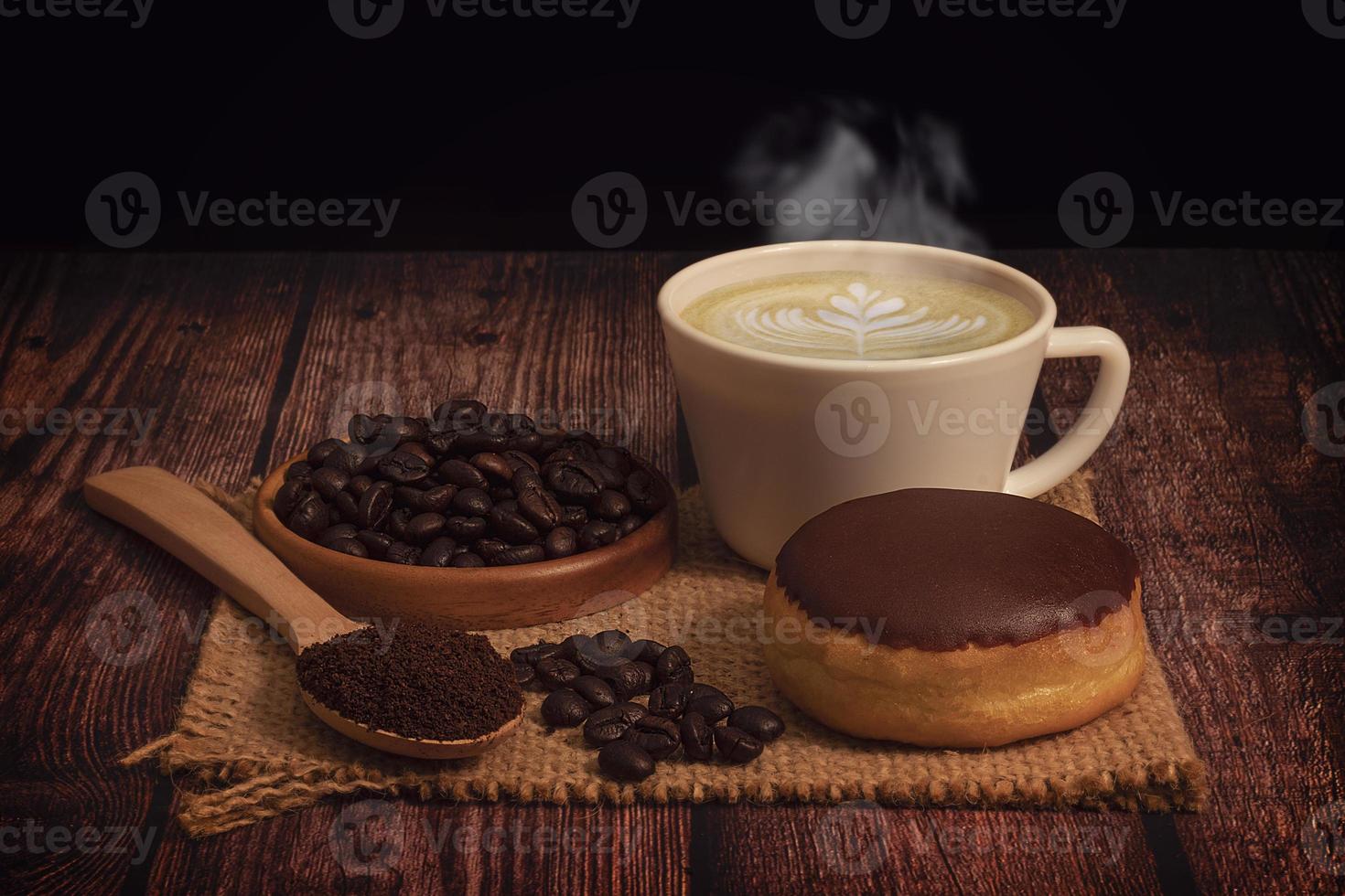 Donut, Tasse dampfenden Kaffee mit Latte Art und Kaffeebohnen auf Sackleinenmatte auf einem Holztisch und schwarzem Hintergrund foto
