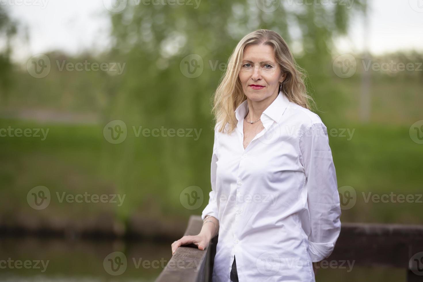 ein schön mittleren Alters europäisch Frau. fünfzig Jahre alt blond Frau im ein Weiß Hemd auf das Hintergrund von Natur. foto