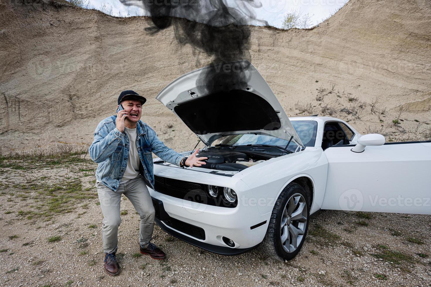 schockierter mann, der phoen zum notfall in der nähe seines kaputten muskelautos ruft, offene motorhaube, rauch vom motor, zusammenbruch, brauche hilfe. foto