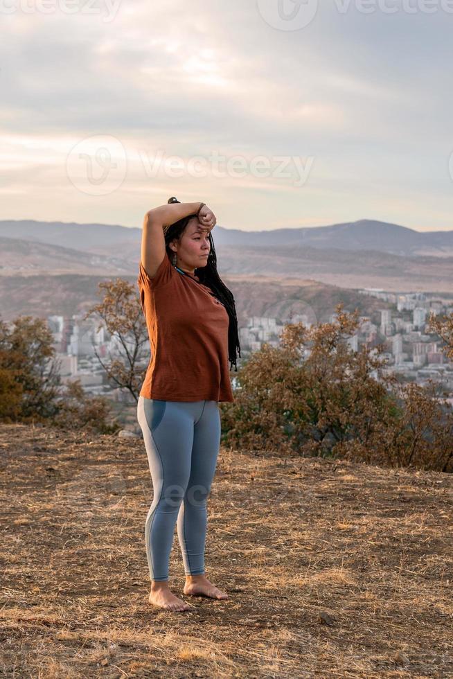 Porträt von schön europäisch Frau mit lange afrikanisch Zöpfe. Frau ist tun Yoga Übung draußen auf Hintergrund von Berge. foto