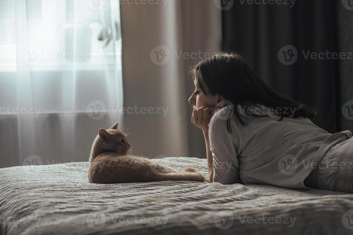 Ein schönes junges Mädchen im Schlafanzug liegt zu Hause auf einem Bett neben ihrer pfirsichfarbenen britischen Katze foto