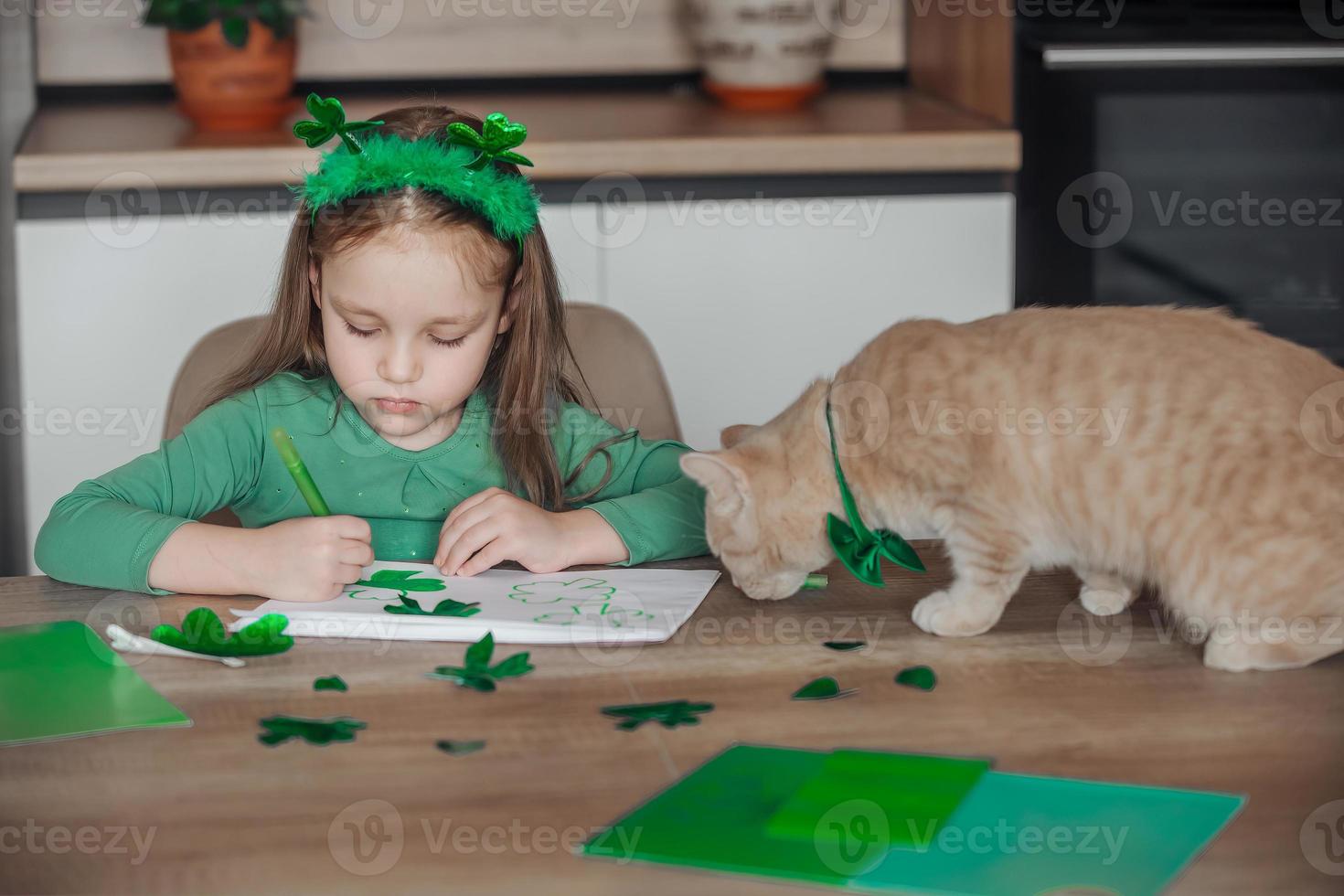 ein wenig Mädchen mit ein Binde auf ihr Kopf zeichnet und schneidet Grün Kleeblätter zum st. Patrick's Tag beim ein Tabelle beim Zuhause im das Küche, Nächster zu ihr ist ihr schön Katze mit ein Grün Bogen Krawatte um seine Hals foto