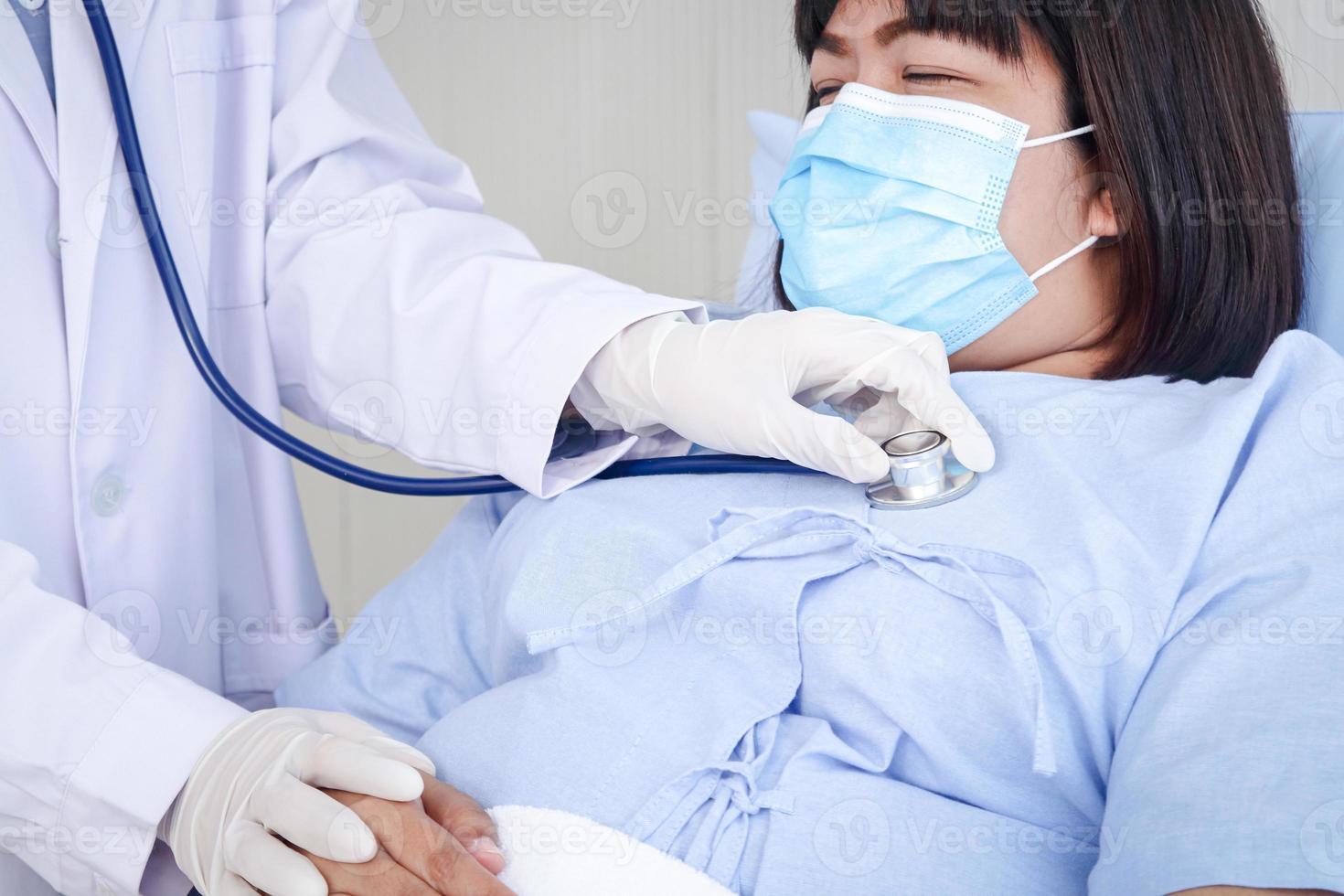 Ärzte benutzt ein Stethoskop zu untersuchen das Gesundheit von ein Fett Frau, tragen ein Maske, Lügen auf ein Krankenhaus Bett. Behandlung von Patienten während das Coronavirus Epidemie. Konzept von medizinisch Bedienung foto