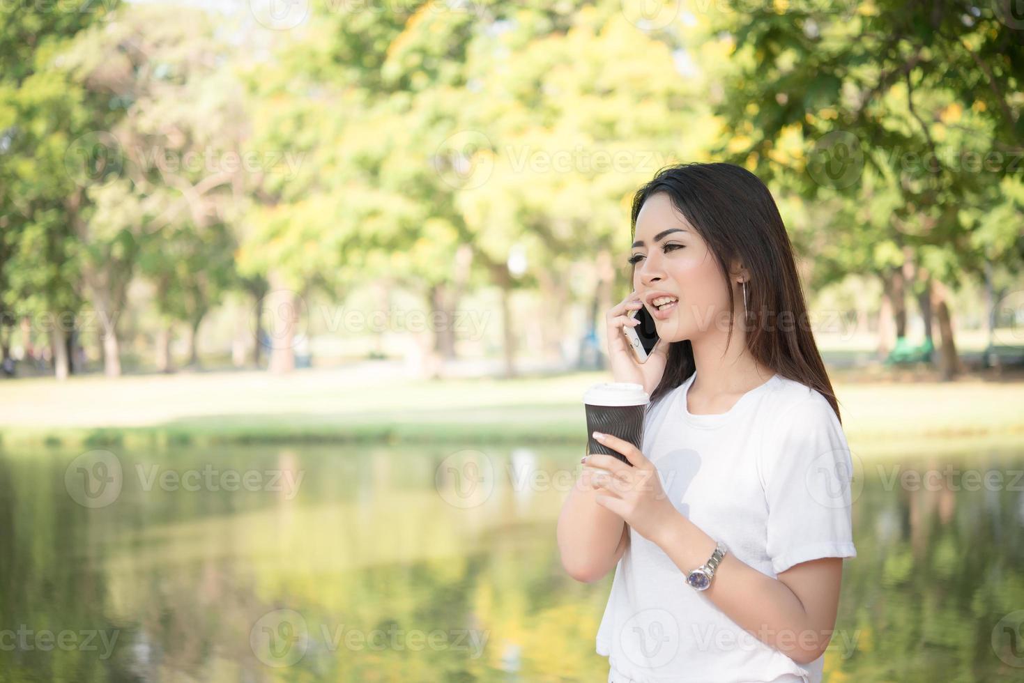 junge schöne Frau, die wegwerfbare Kaffeetasse hält, während sie am Telefon spricht foto