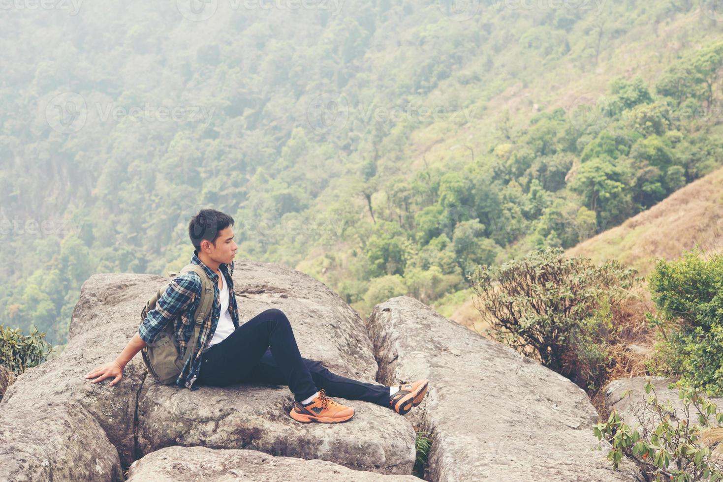 junger Hipster-Wanderer mit Rucksack, der oben auf dem Berg sitzt foto