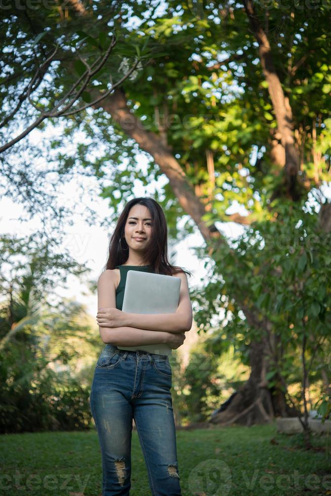 junge schöne Frau, die einen Laptop-Computer hält, während an einem Park entspannen geht foto