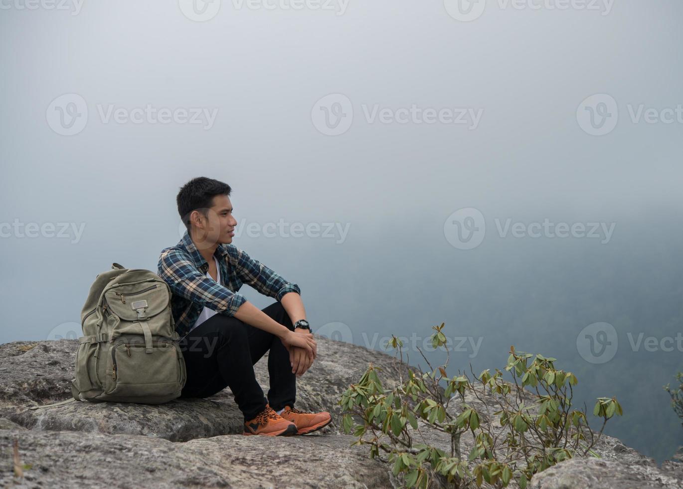 junger Hipster-Wanderer mit Rucksack, der oben auf dem Berg sitzt foto