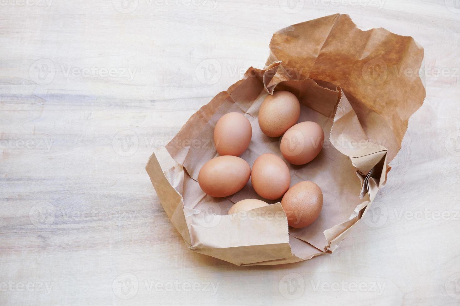 braunes Ei in Papiertüte dahinter, frische Hühnereier auf Holztisch foto